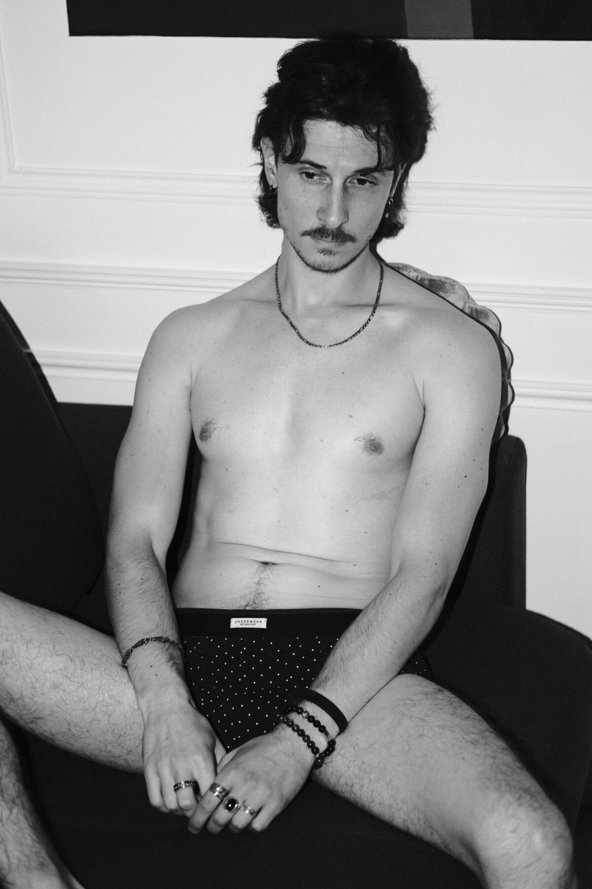 Portrait noir et blanc de Nico assis en sous-vêtement noir à pois, torse nu, bagues aux mains, regard vers l'objectif