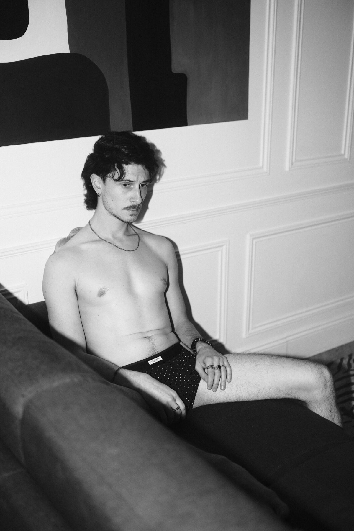 Portrait noir et blanc de Nico assis en sous-vêtement noir à pois, jambe tendue, bracelet et bagues, regard baissé