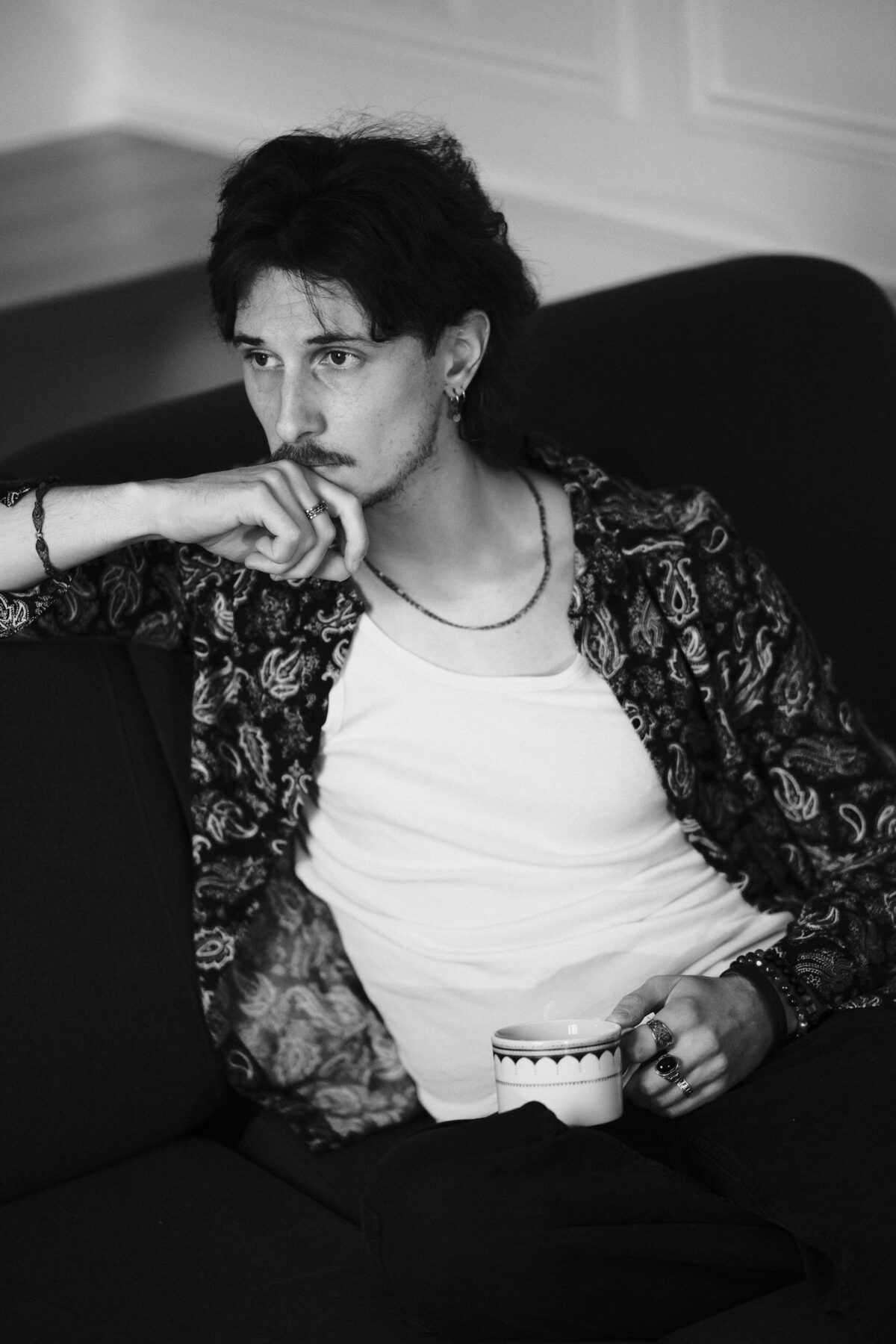 Portrait noir et blanc de Nico assis sur un canapé, chemise paisley ouverte, main sous le menton ornée de bagues, tasse à la main
