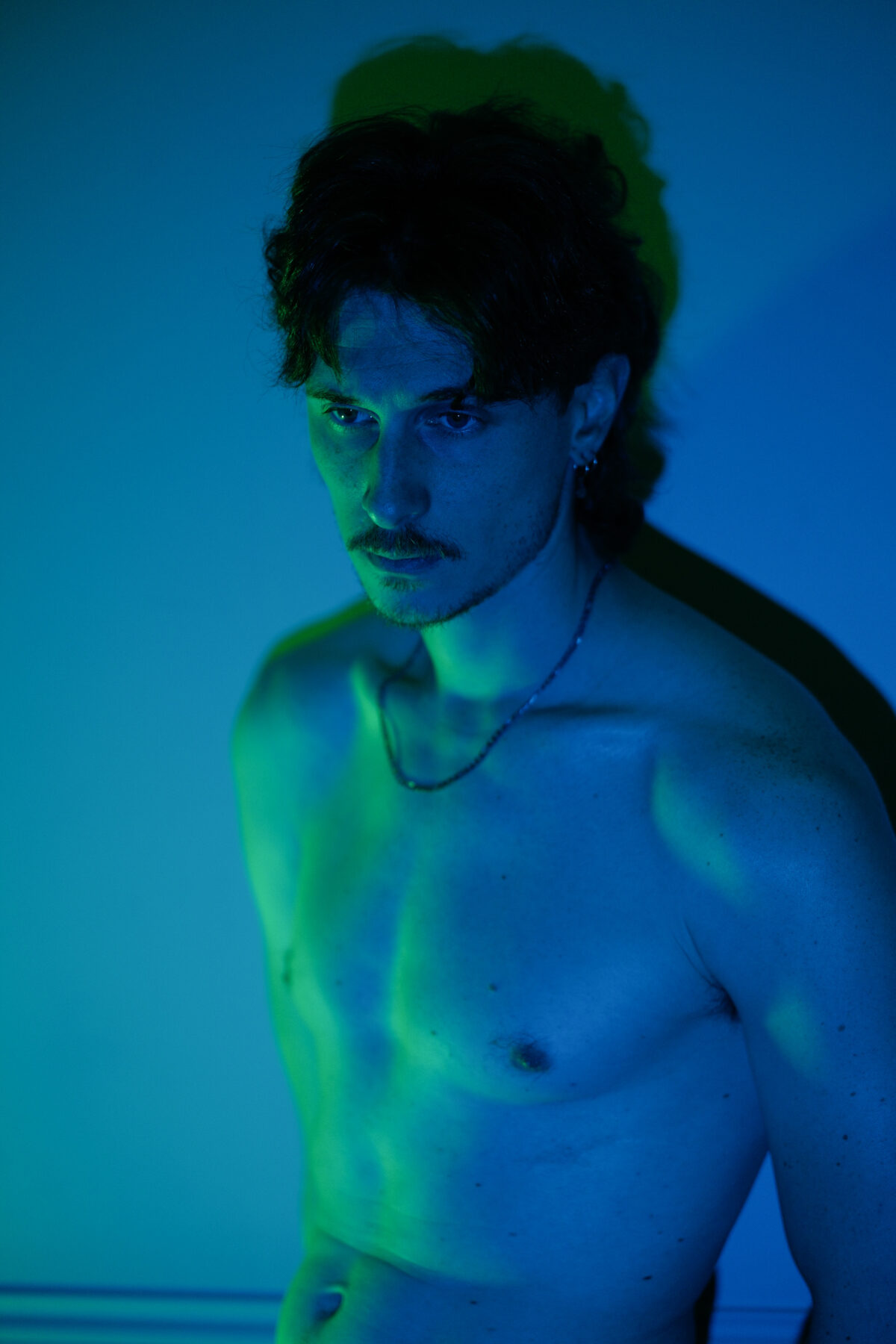 Portrait de Nico torse nu sous une lumière bleue et verte, fine moustache, collier au cou, regard baissé