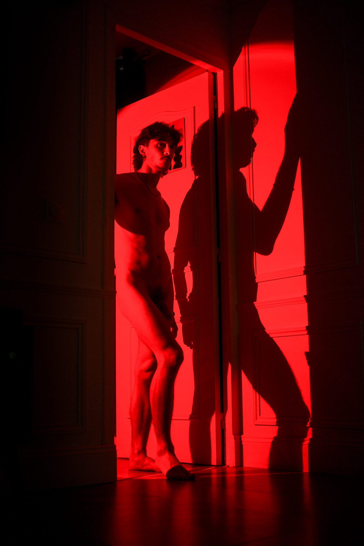 Silhouette nue de Nico dans l'embrasure d'une porte sous une lumière rouge, ombre projetée sur le mur, regard vers l'objectif
