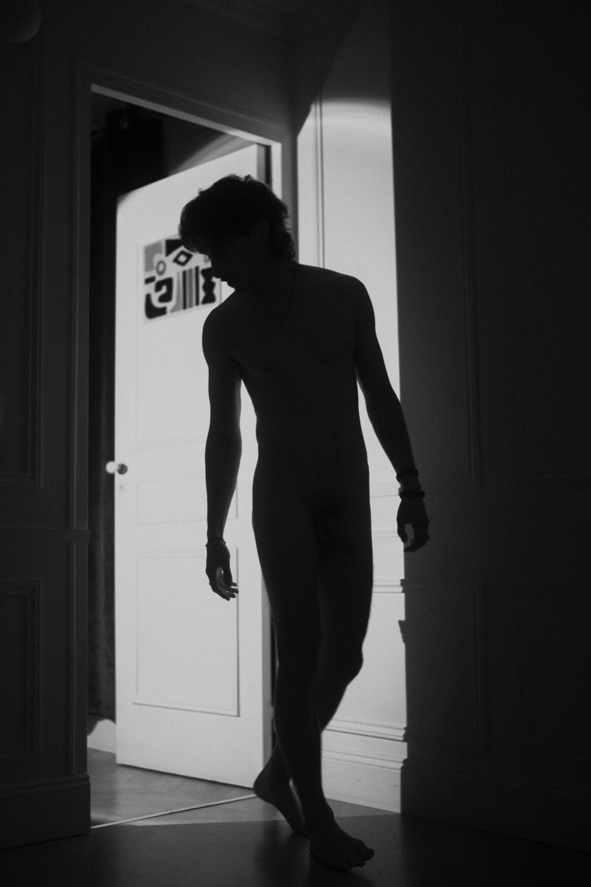 Silhouette nue et en noir et blanc de Nico s'avançant dans l'embrasure d'une porte éclairée, tête baissée, chevelure bouclée