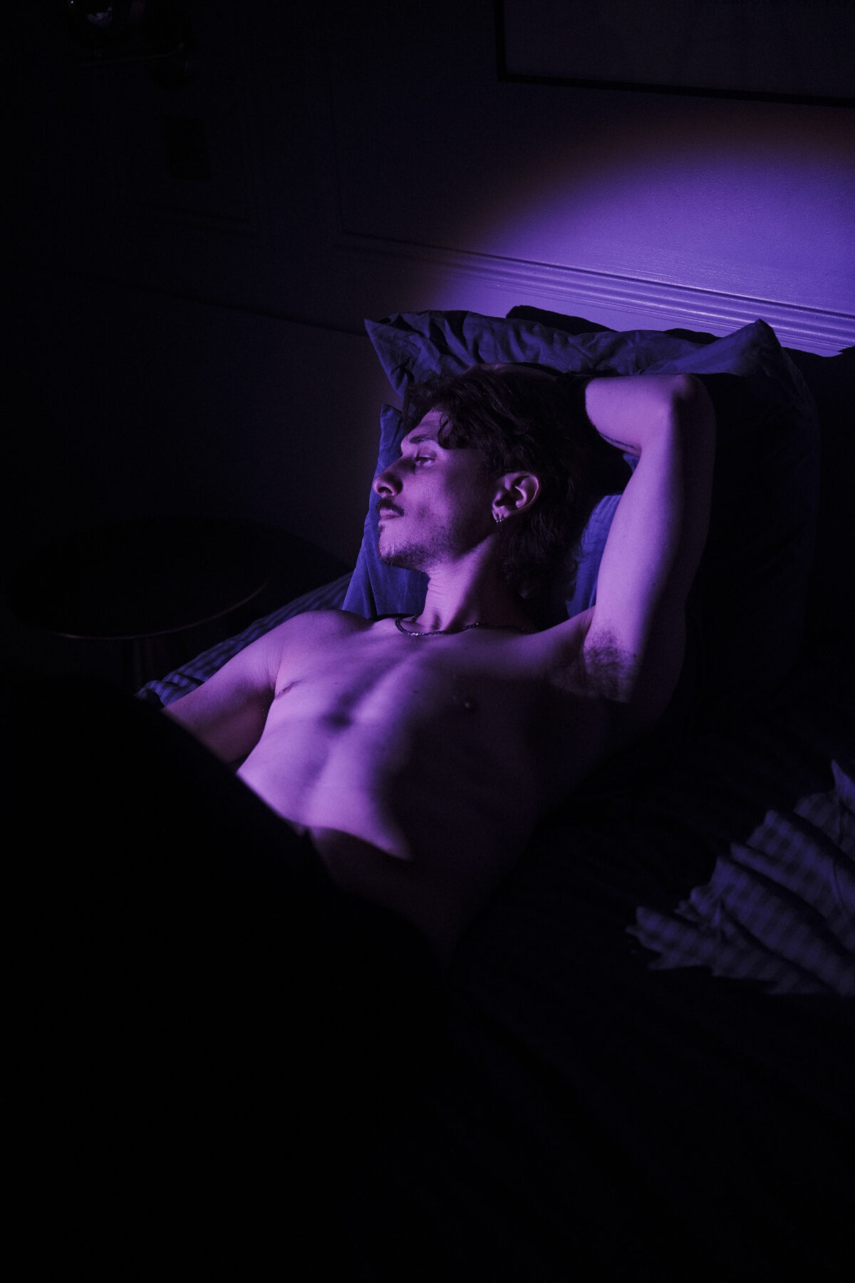 Portrait de Nico allongé, torse nu, éclairage violet dramatique, bras replié sous la tête
