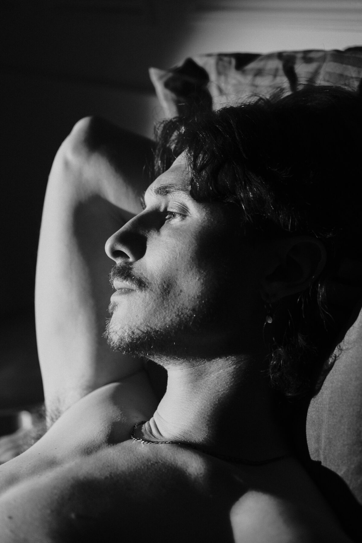 Portrait noir et blanc de profil de Nico allongé, bras levé, collier fin, lumière naturelle contrastée