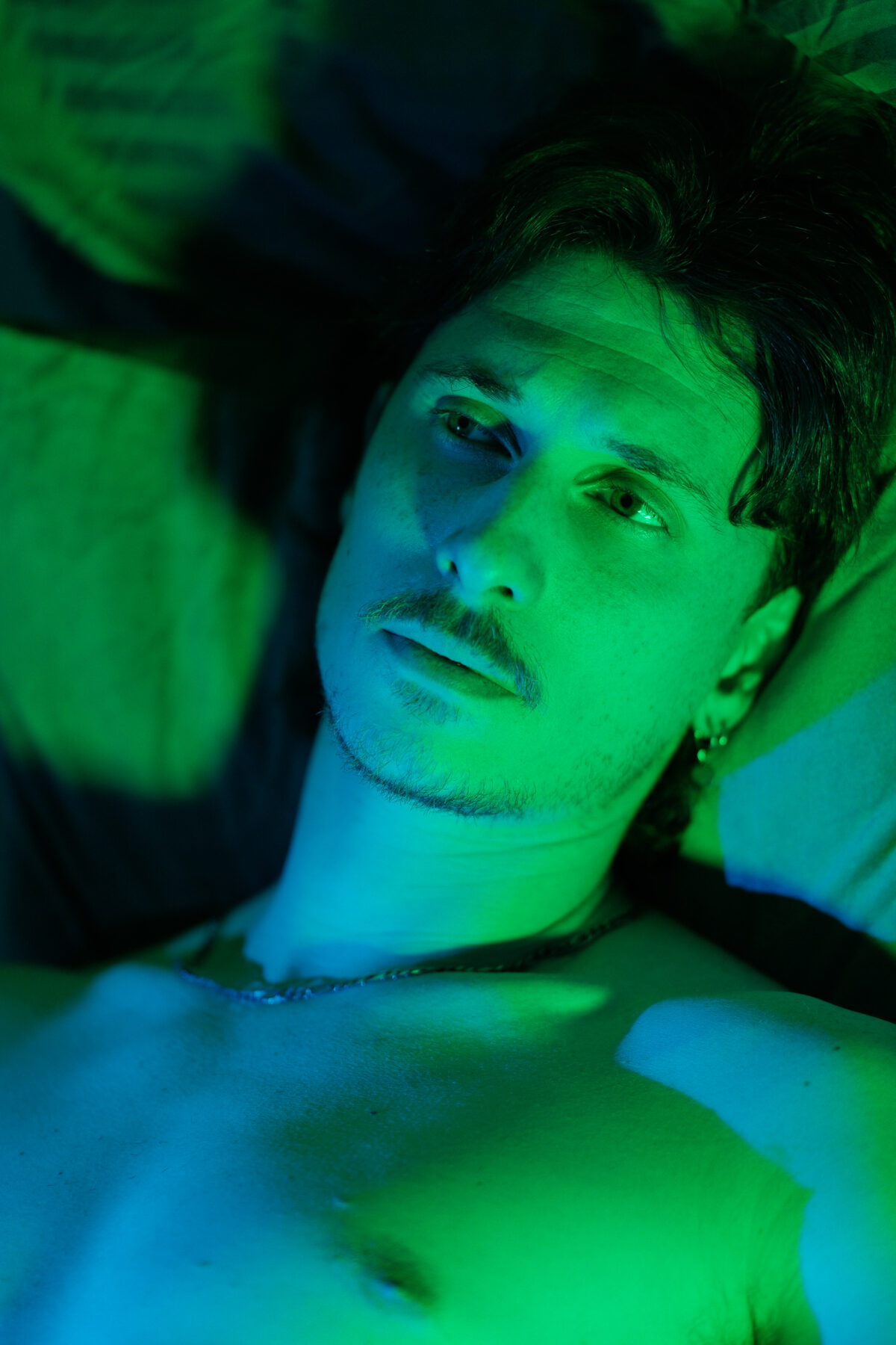 Portrait de Nico allongé, torse nu, éclairage vert et bleu, regard de côté, boucle d'oreille visible