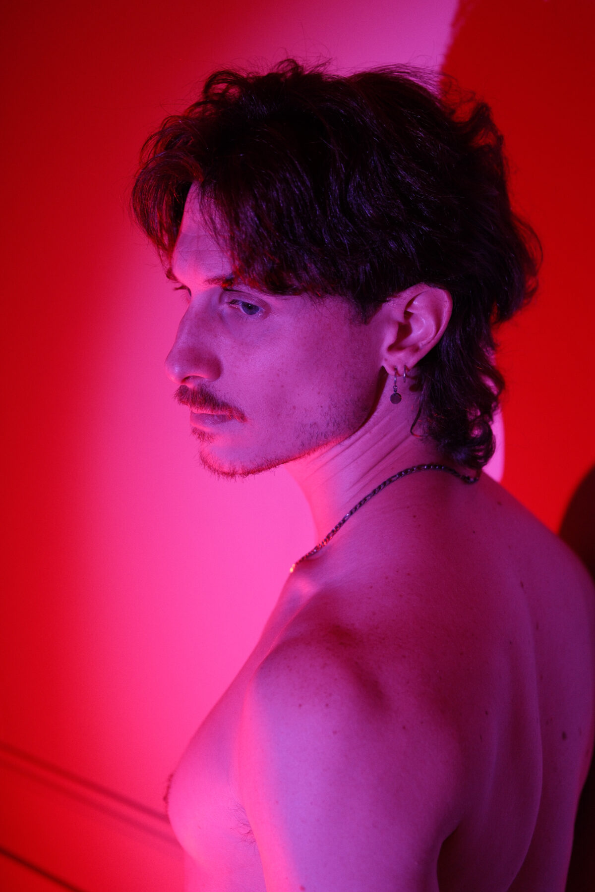 Portrait de profil de Nico, torse nu, éclairage rouge et rose, boucle d'oreille et collier visibles