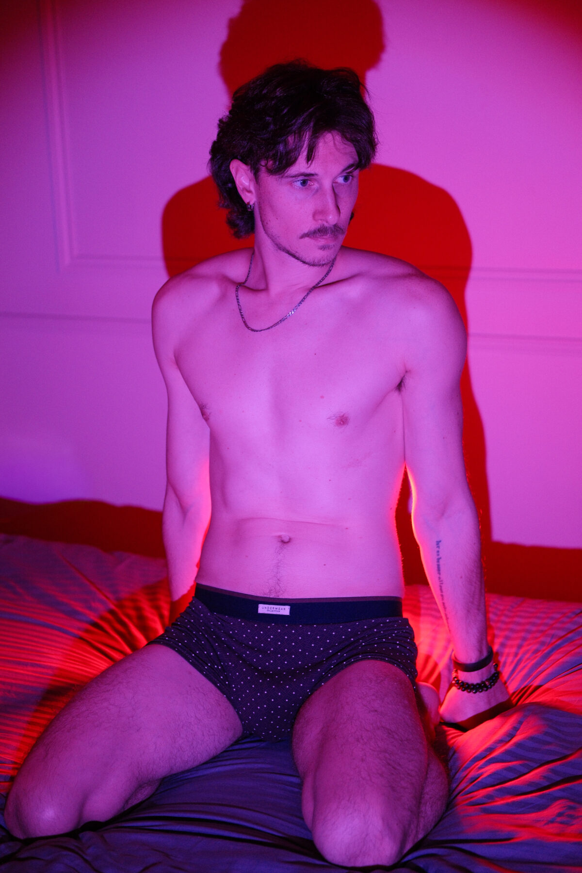 Portrait de Nico agenouillé sur un lit en sous-vêtement noir à pois, tatouage sur l'avant-bras, éclairage rose et violet