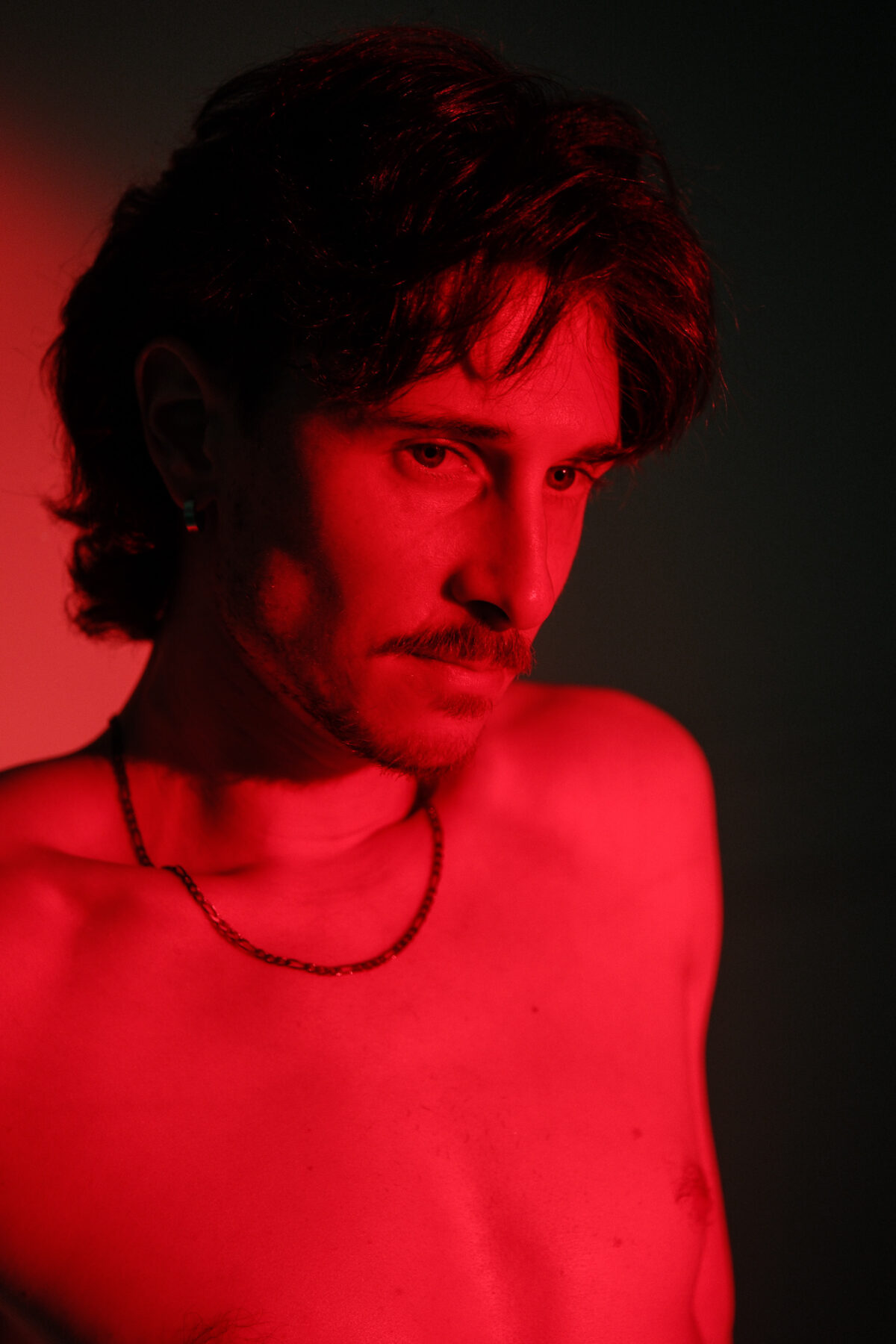 Portrait rapproché de Nico sous éclairage rouge, collier fin, regard baissé, ambiance dramatique