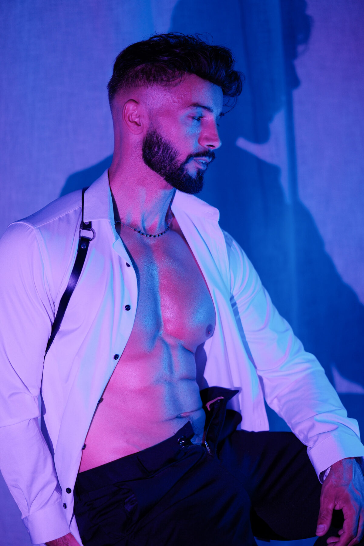 Andy en chemise blanche ouverte et harnais en cuir, torse musclé, regard de côté, lumière bleue et violette