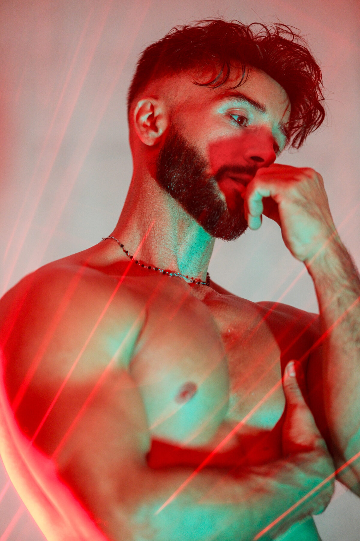 Andy, main près de la bouche, torse nu et reflets laser rouges sur fond clair