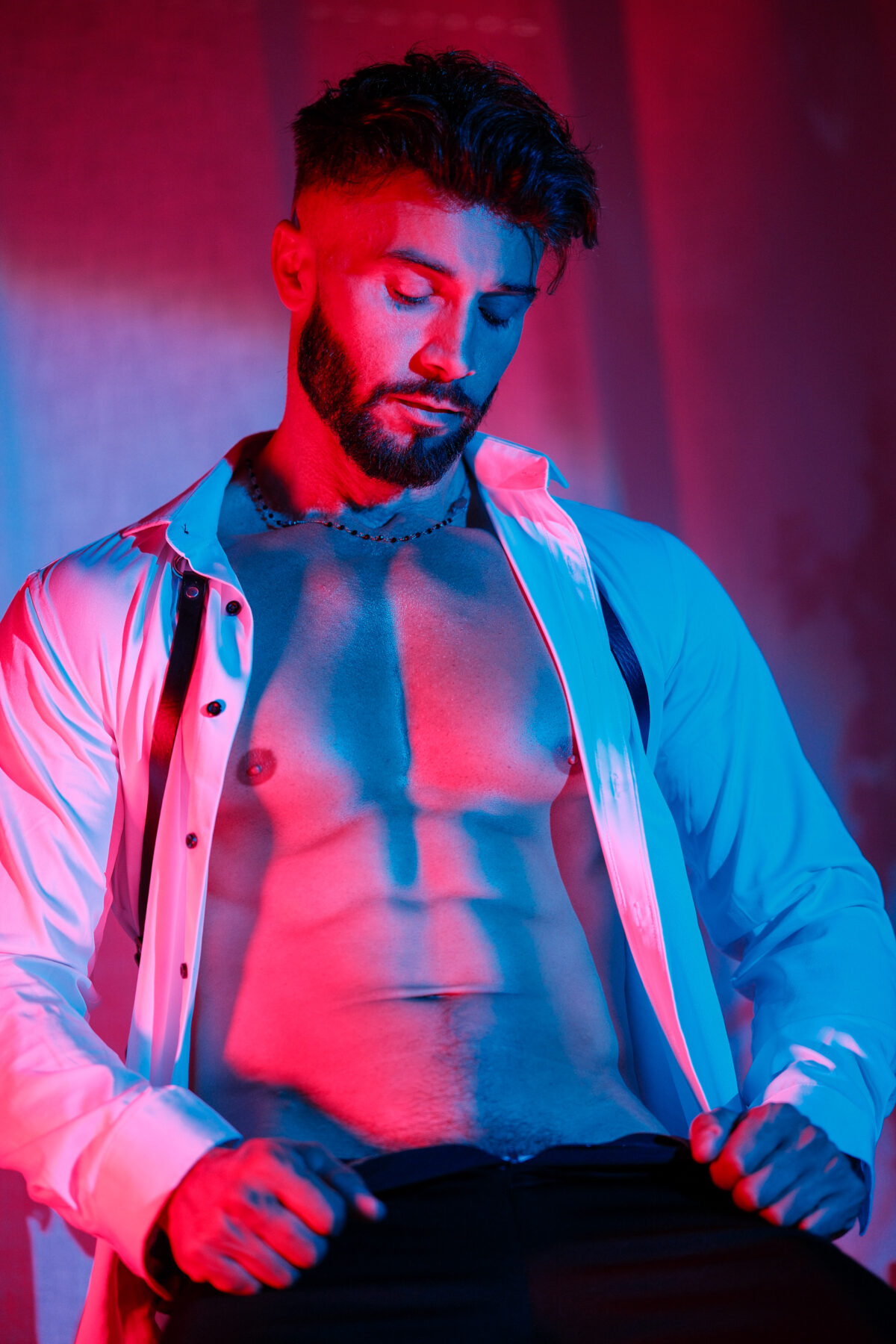Andy tête baissée, chemise blanche ouverte sur un torse musclé, lumière rouge et bleue