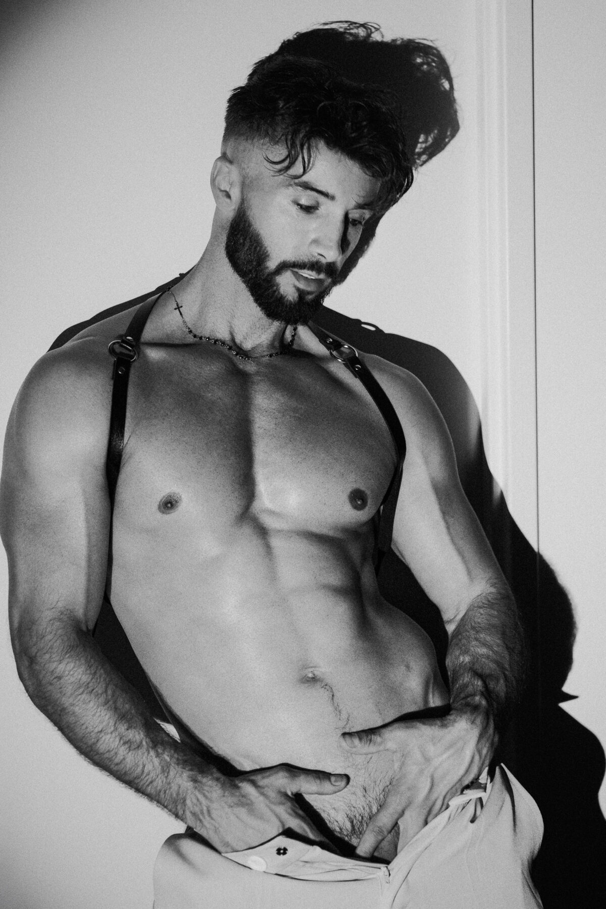 Andy en noir et blanc, harnais et pantalon déboutonné, main sur le ventre, regard baissé