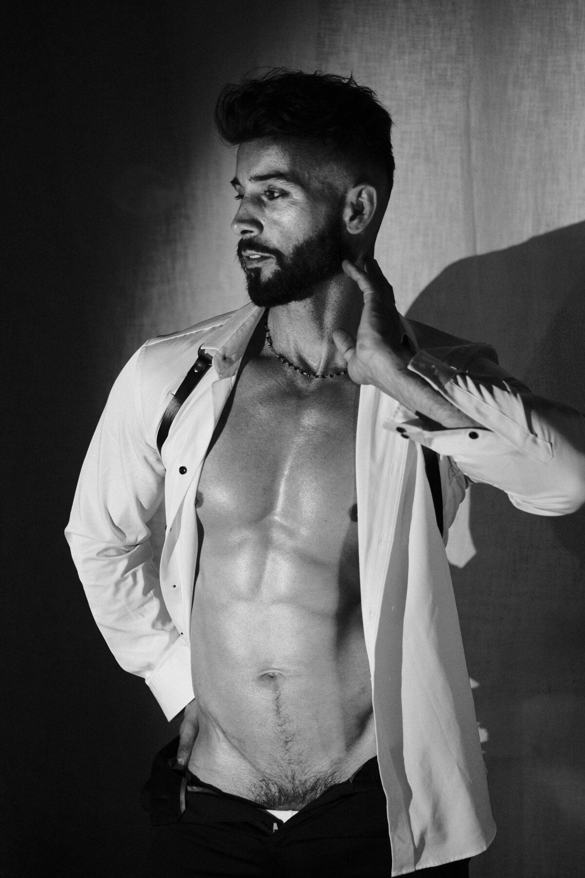 Portrait noir et blanc d'Andy, chemise blanche ouverte et harnais, main sur la nuque, torse sculpté