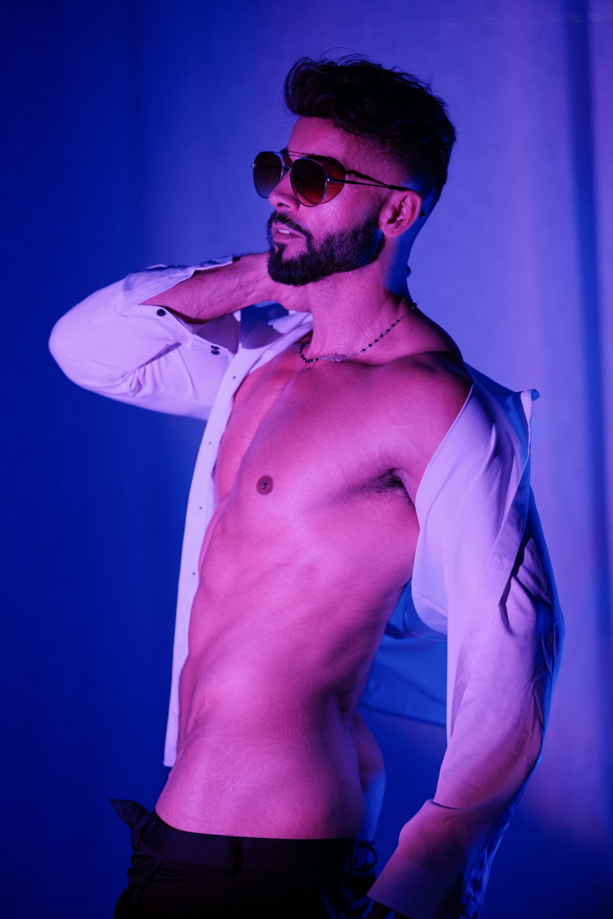 Andy en lunettes de soleil aviateur, chemise blanche glissant des épaules, torse nu en lumière violette