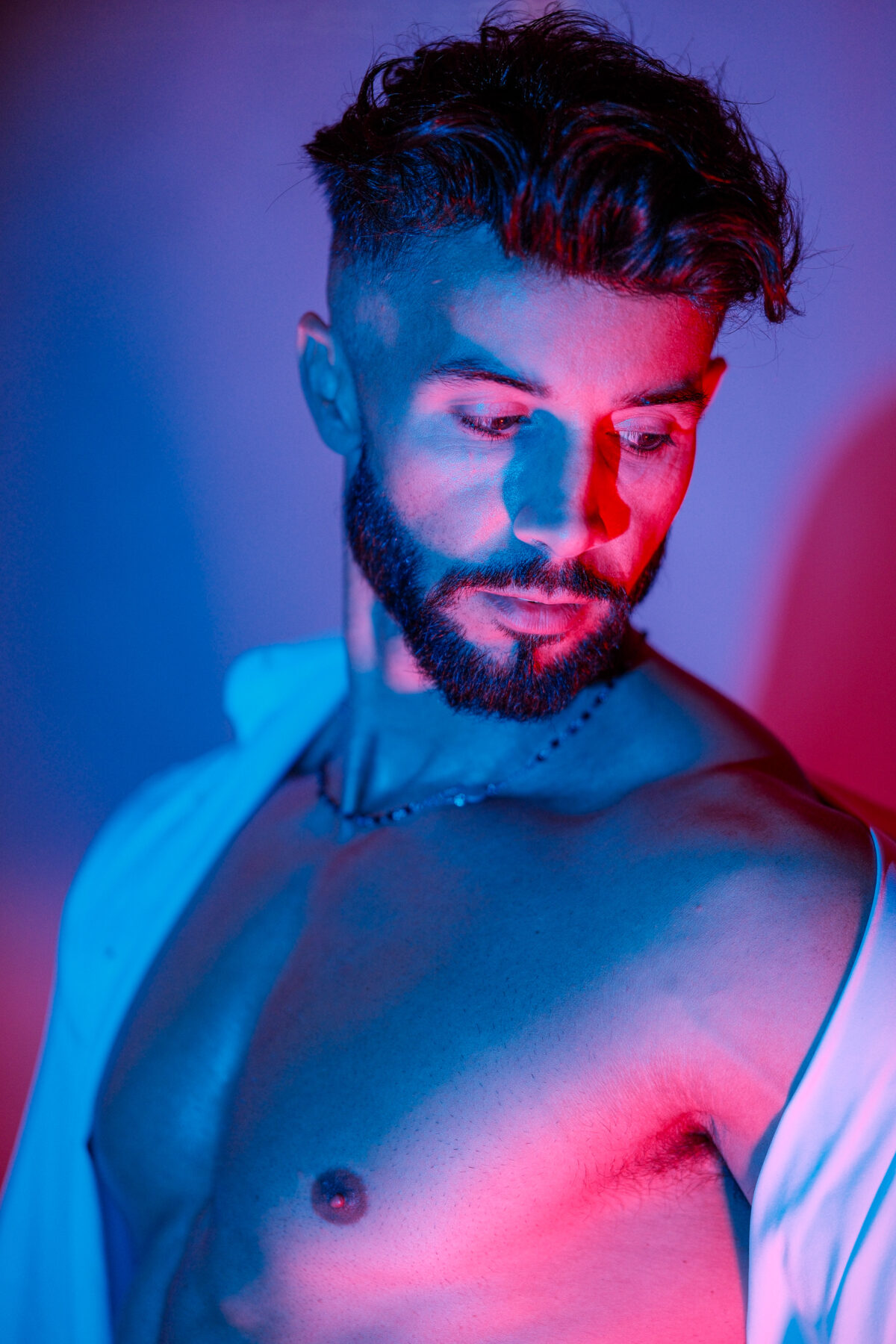 Portrait d'Andy, barbe fournie et regard baissé, épaule nue sous une lumière bleue et rouge