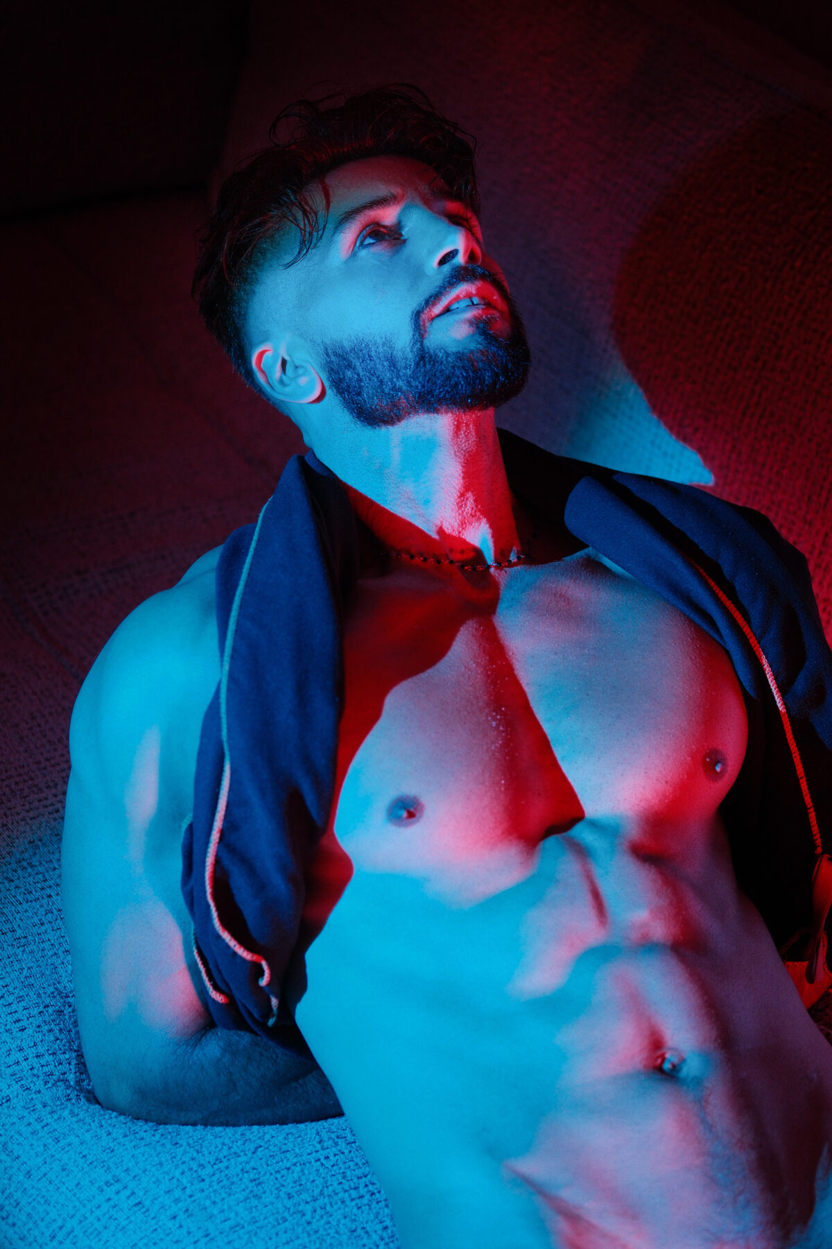 Andy allongé, photographié en contre-plongée, veste sombre ouverte sur un torse musclé