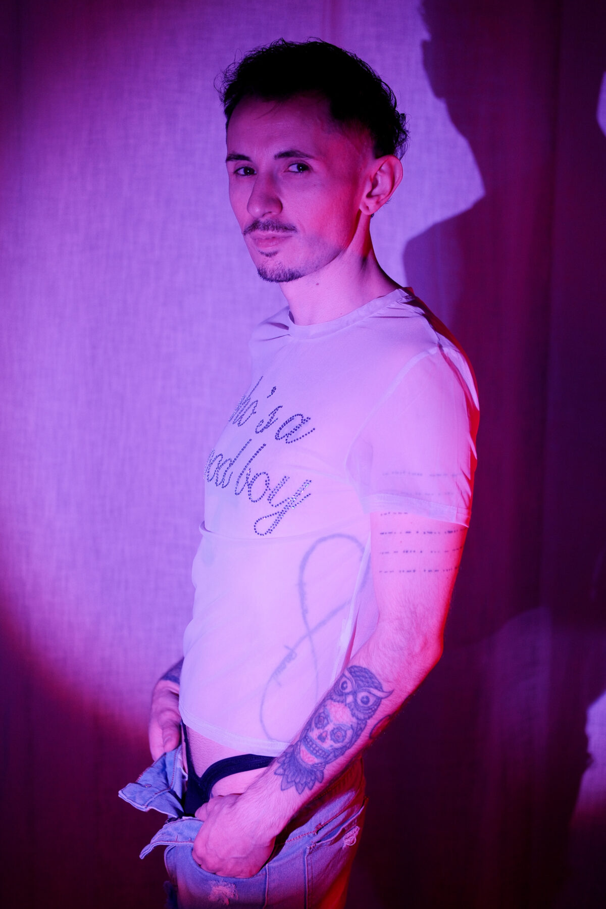 Romain debout de profil, T-shirt transparent à strass, mains dans son short en jean, lumière violette