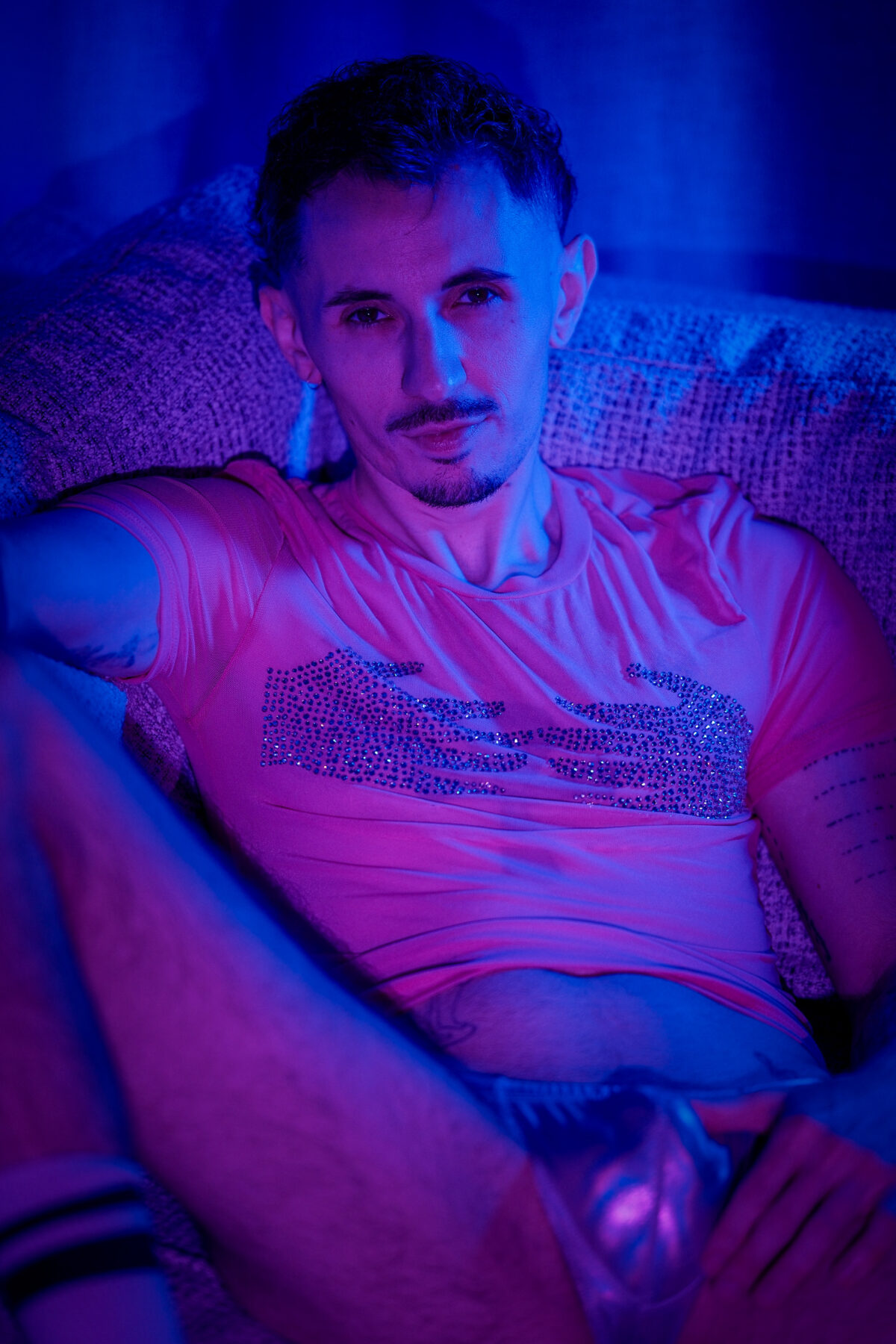Romain assis sur un canapé, T-shirt rose à strass, lumière bleue et rose, regard direct