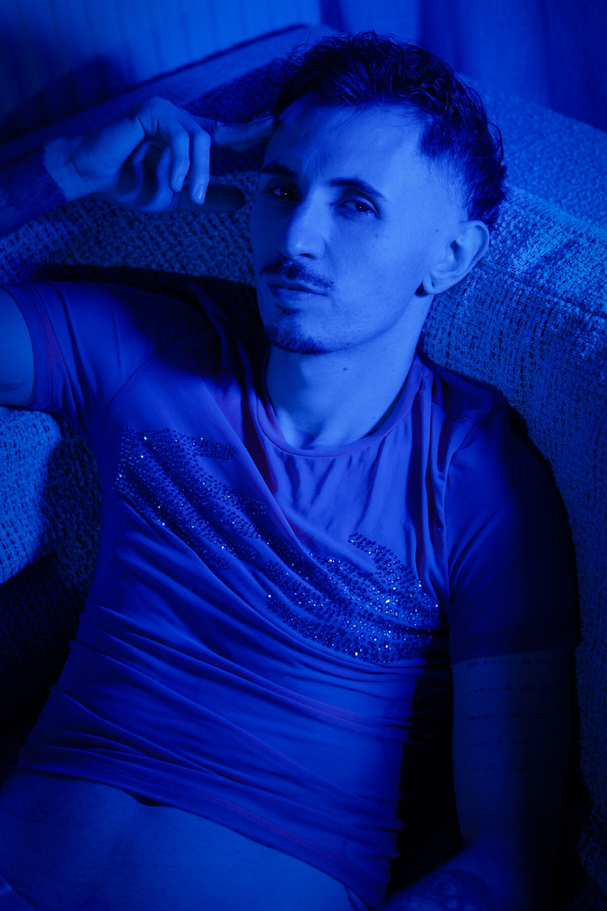Portrait homme de Romain en lumière bleue, main sur la tempe, allongé sur un canapé, T-shirt à strass