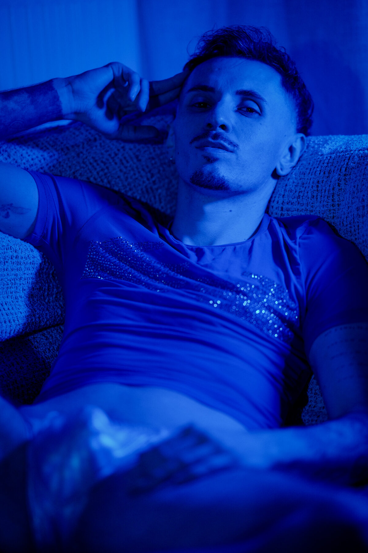 Romain allongé sur un canapé en lumière bleue, main derrière la tête, regard intense, T-shirt à strass
