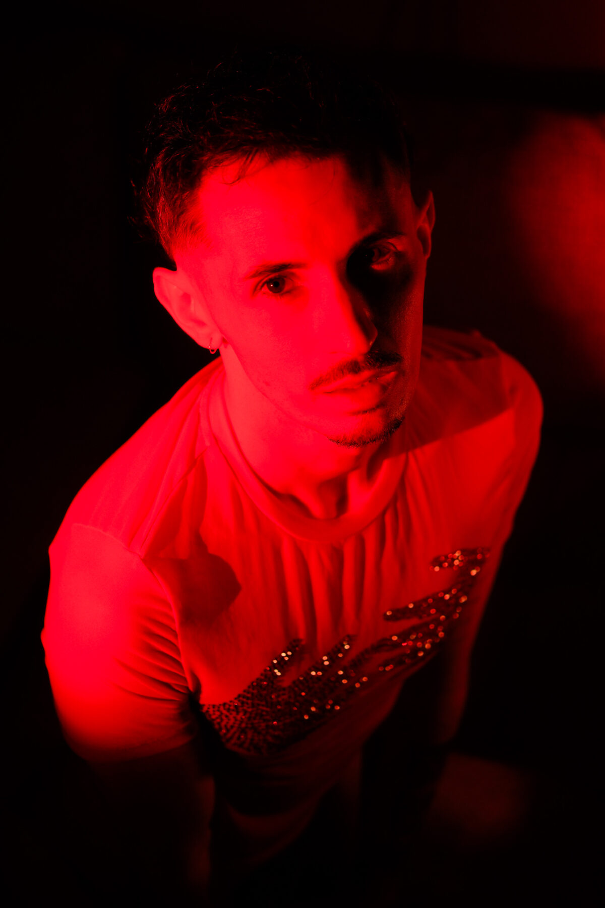 Portrait homme de Romain en plongée, éclairage rouge sur fond noir, T-shirt à strass, regard franc