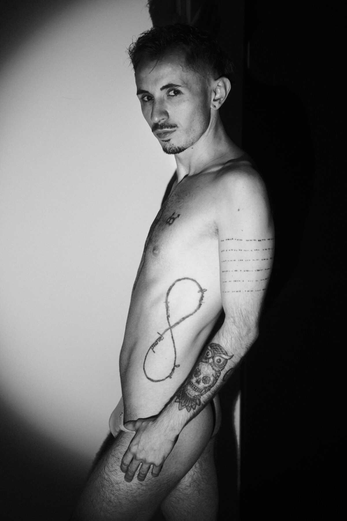 Portrait noir et blanc de Romain de profil, torse nu tatoué, symbole infini sur les côtes, halo de lumière