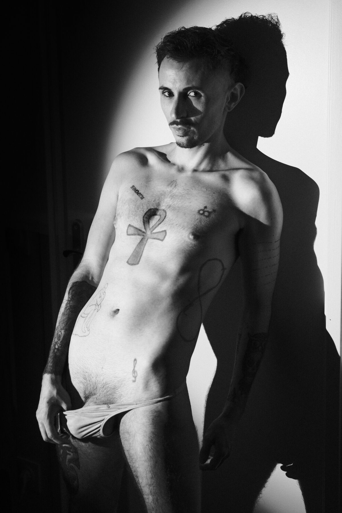 Romain torse nu en noir et blanc, tatouage ankh sur le torse, ombre portée sur le mur, main sur son slip