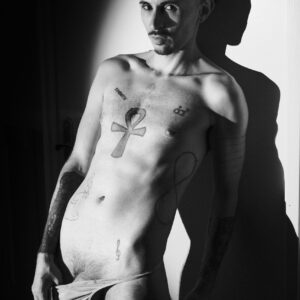 Romain torse nu en noir et blanc, tatouage ankh sur le torse, ombre portée sur le mur, main sur son slip