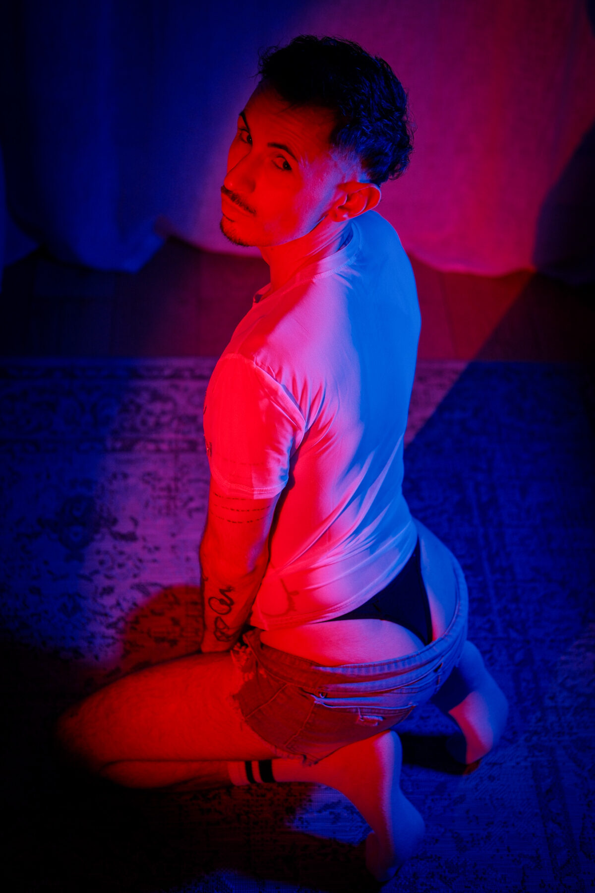 Romain de dos, à genoux, regard par-dessus l'épaule, T-shirt blanc sous une lumière rouge et bleue