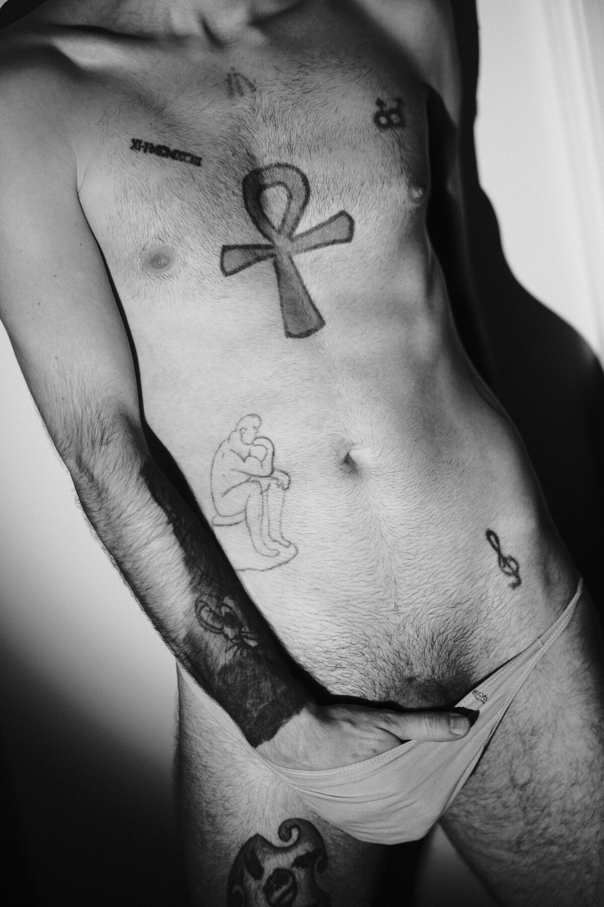 Gros plan noir et blanc sur le torse tatoué de Romain, ankh et clé de sol, main dans son slip
