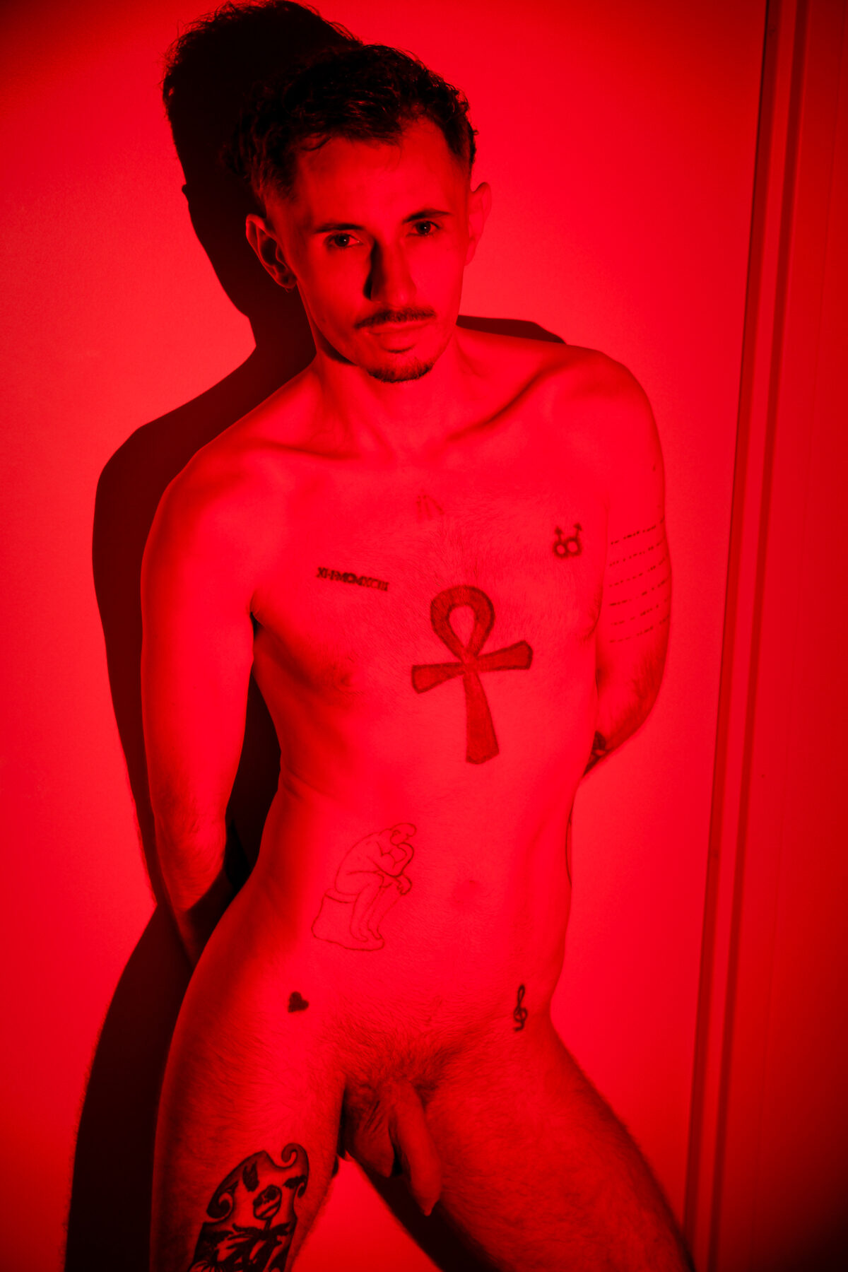Nu artistique de Romain debout en lumière rouge, mains dans le dos, tatouage ankh sur le torse