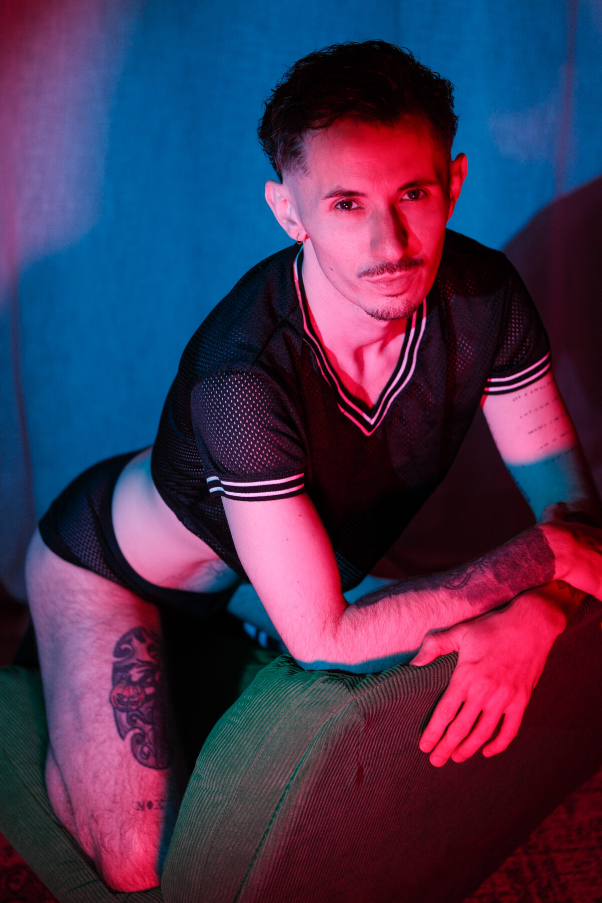 Romain à genoux, appuyé sur un pouf, T-shirt résille noir, lumière bleue et rouge, cuisse tatouée