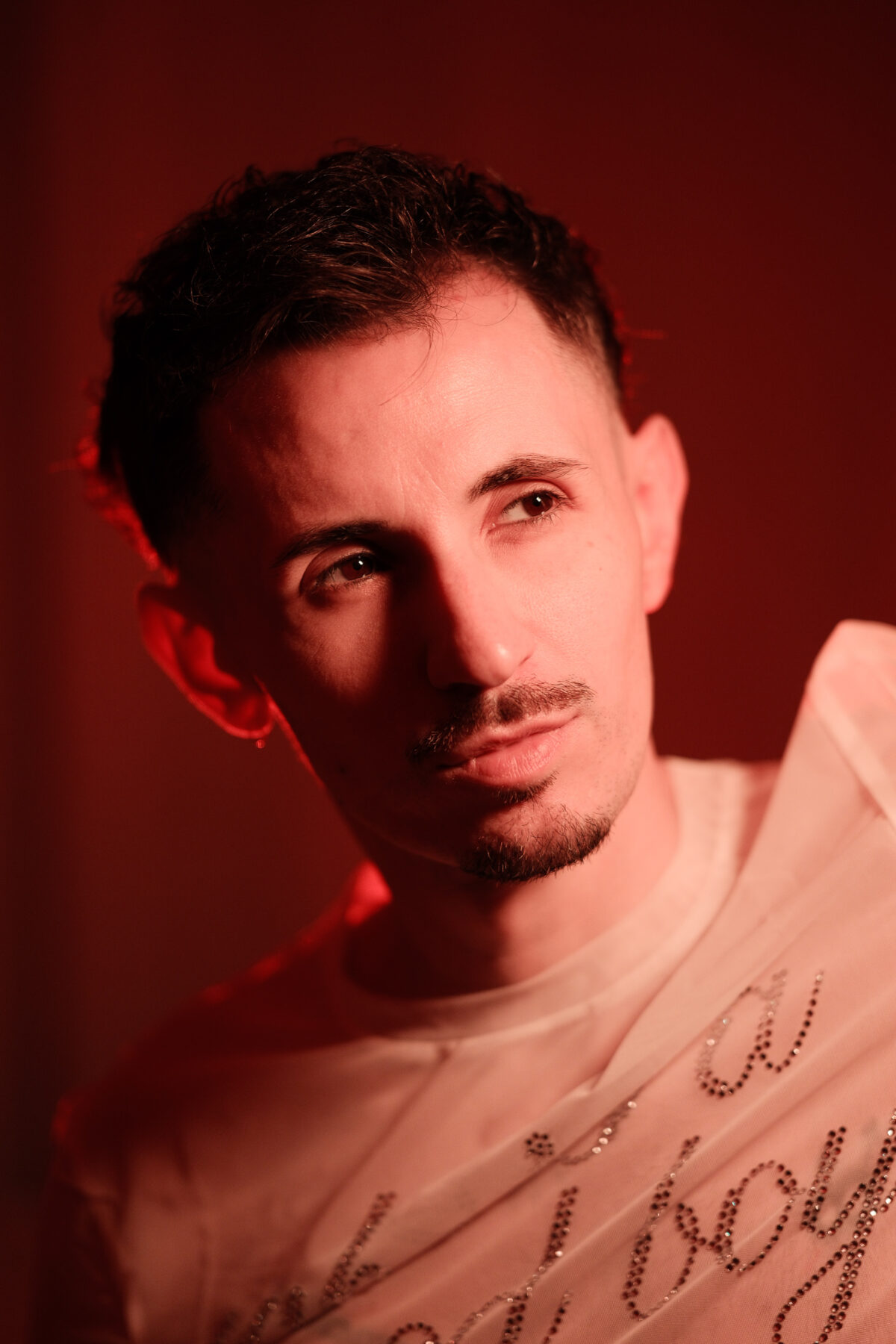 Portrait homme de Romain en lumière rouge, regard de côté, moustache et barbiche, T-shirt blanc à strass